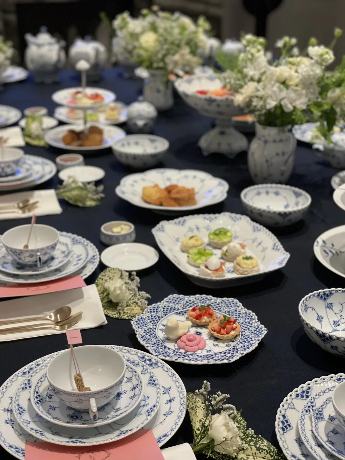 생일 티클래스 남색 테이블보 위에 찻잔 세트와 다과 접시, 흰 꽃 장식이 길게 놓인 테이블을 위에서 본 장면